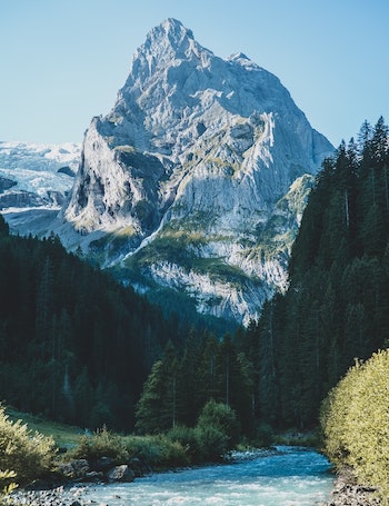a jagged, snowy mountain with trees and a river in the foreground
